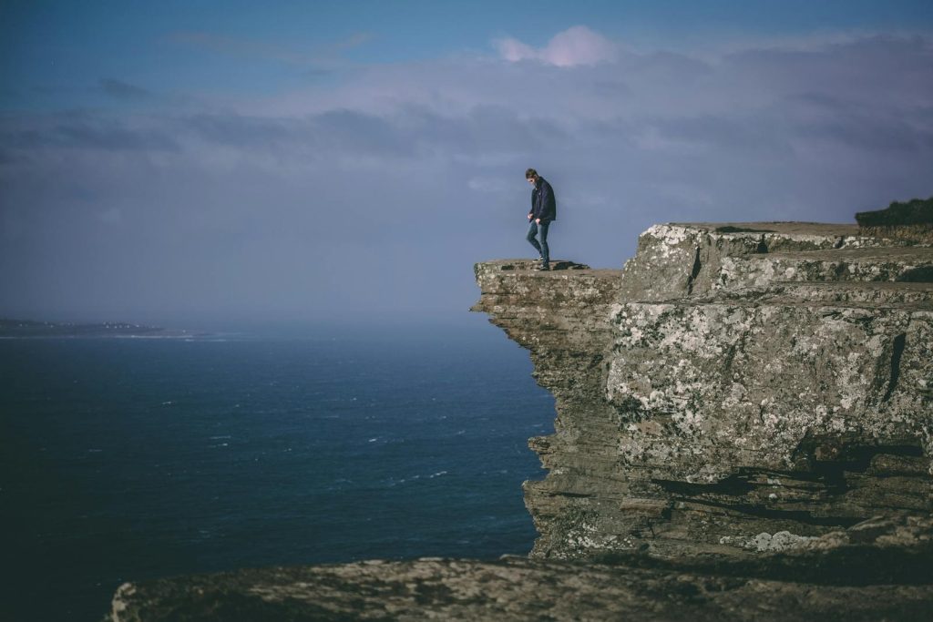Courageous individual standing atop a rocky cliff with ocean view, embracing uncertainty, challenge, and freedom over the false security of a paycheck