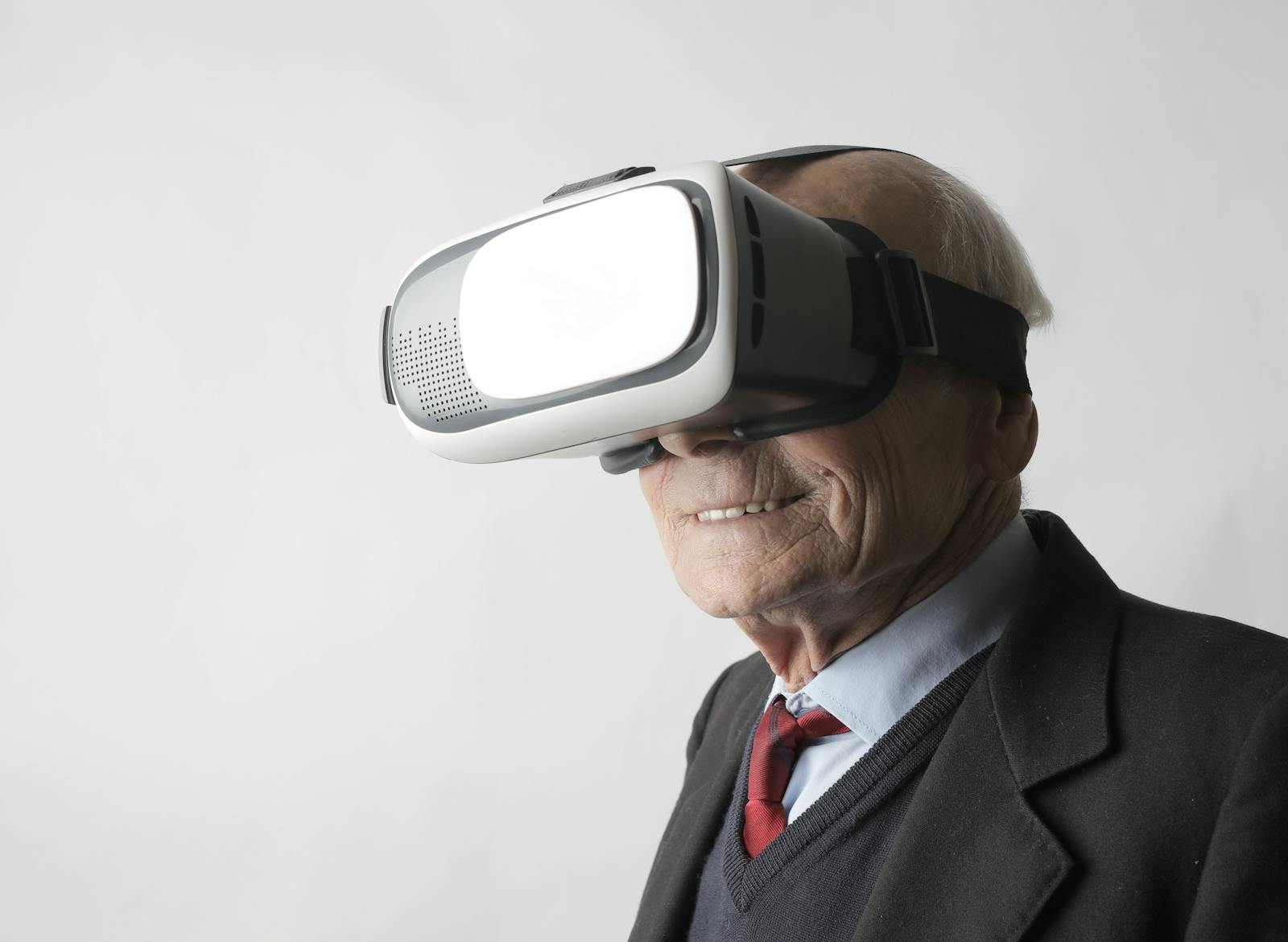 Smiling elderly gentleman wearing classy suit experiencing virtual reality while using modern headset on white background.